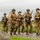 WWII Reenactors Marching at the at the LT John S Thompson Bridge Ceremony, Operation Market Garden Commemorations 2026 A group of individuals dressed in WWII-era U.S. Army uniforms are marching along a rural road, carrying period-accurate rifles. The uniforms feature American flag patches on the sleeves, M1 steel helmets, and olive drab fatigues consistent with mid-20th century U.S. military dress. The setting and well-preserved costumes suggest this is a historical reenactment event.