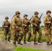 A group of individuals dressed in WWII-era U.S. Army uniforms are marching along a rural road, carrying period-accurate rifles. The uniforms feature American flag patches on the sleeves, M1 steel helmets, and olive drab fatigues consistent with mid-20th century U.S. military dress. The setting and well-preserved costumes suggest this is a historical reenactment event.