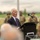 Dutch Survivor Remembers his Childhood Experience During Operation Market Garden, Operation Market Garden Commemorations 2026 An elderly man in a dark suit and purple tie is delivering a speech at a podium with a microphone at what appears to be a military ceremony or commemoration event. Several soldiers in camouflage uniforms are standing at attention in the blurred background. The setting appears to be an outdoor venue with green fields visible behind the assembled personnel.