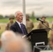 An elderly man in a dark suit and purple tie is delivering a speech at a podium with a microphone at what appears to be a military ceremony or commemoration event. Several soldiers in camouflage uniforms are standing at attention in the blurred background. The setting appears to be an outdoor venue with green fields visible behind the assembled personnel.