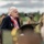 Dutch Survivor reads Poetry Remembering the Allied Sacrifices During Operation Market Garden, Operation Market Garden Commemorations 2026 An elderly woman with white hair is speaking at a podium with a microphone at what appears to be a military ceremony or commemoration event. She is wearing a dark navy blazer with military unit patches on the lapel and a red, white, and blue scarf. Military personnel in uniform are visible in the blurred background.