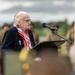 An elderly woman with white hair is speaking at a podium with a microphone at what appears to be a military ceremony or commemoration event. She is wearing a dark navy blazer with military unit patches on the lapel and a red, white, and blue scarf. Military personnel in uniform are visible in the blurred background.