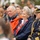 Ms. Chloe Gavin Reacts During a Speech at the LT John S Thompson Bridge Ceremony, Operation Market Garden Commemorations 2026 A group of people are seated at what appears to be a formal outdoor ceremony or commemorative event. The foreground features an elderly couple and a senior naval officer in full dress uniform with gold braid and medals. The woman is wearing a scarf with what appears to be Norwegian flag colors, suggesting a Norwegian national or military event.