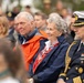 A group of people are seated at what appears to be a formal outdoor ceremony or commemorative event. The foreground features an elderly couple and a senior naval officer in full dress uniform with gold braid and medals. The woman is wearing a scarf with what appears to be Norwegian flag colors, suggesting a Norwegian national or military event.