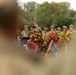A military officer in dress uniform delivers a speech at a podium during what appears to be a memorial or commemorative ceremony. Multiple floral wreaths with colored ribbons are prominently displayed, and soldiers wearing WWII-era helmets stand in formation in the background. The scene suggests a formal remembrance event, likely honoring fallen soldiers.