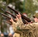 A group of individuals dressed in WWII-era U.S. Army uniforms are performing a rifle salute or ceremonial volley fire. They are firing M1 Garand semi-automatic rifles upward at an angle, consistent with a military honors ceremony. A spent brass casing is visibly ejecting from the nearest rifle, indicating live or blank fire.