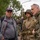 Attendees at the LT John S Thompson Bridge Ceremony, Operation Market Garden Commemorations 2026 Three men are gathered at what appears to be a WWII commemorative event or reenactment. Two of the men are wearing 82nd Airborne Division insignia — one in period-accurate WWII U.S. Army uniform with helmet and field gear, and another in an olive drab jacket with an 82nd Airborne patch. The setting appears to be an outdoor public gathering with other attendees visible in the background.