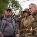 Three men are gathered at what appears to be a WWII commemorative event or reenactment. Two of the men are wearing 82nd Airborne Division insignia — one in period-accurate WWII U.S. Army uniform with helmet and field gear, and another in an olive drab jacket with an 82nd Airborne patch. The setting appears to be an outdoor public gathering with other attendees visible in the background.