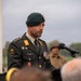 A military officer in a dark green dress uniform and beret is speaking at a podium with a microphone, likely at a commemorative or ceremonial outdoor event. He wears multiple medals, unit patches including a '13' designation, and rank insignia consistent with a senior officer. The background includes children, civilians, and individuals in period military attire, suggesting a memorial or historical commemoration ceremony.