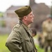 A man dressed in WWII-era U.S. Army olive drab uniform, including a garrison/overseas cap, stands in a field with hands behind his back. He is carrying a modern camera with a leather case on a shoulder strap, suggesting this is a historical reenactment event. Additional figures in period military attire are visible in the blurred background.