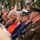 Attendees at the LT John S Thompson Bridge Ceremony, Operation Market Garden Commemorations 2026 A formal military and civilian ceremony is taking place outdoors, with attendees seated in rows facing forward. Two uniformed military officers are prominently visible in the foreground — one in a U.S. Army olive/brown dress uniform and one in a dark naval dress uniform with a white-topped cap. The gathering appears to be a commemorative or memorial event, suggested by the formal attire, flags in the background, and mixed civilian-military attendance.
