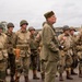A group of individuals dressed in World War II-era U.S. Army uniforms are assembled in formation, likely participating in a historical reenactment or commemorative event. The uniforms, helmets, equipment, and vehicles visible in the background are consistent with American military gear from the 1940s. The setting appears to be an outdoor paved area under overcast skies.