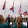 Deputy Mayor of Grave Delivers Remarks, Operation Market Garden Commemorations 2026 An outdoor memorial or commemorative ceremony is taking place, featuring a speaker at a podium wearing a ceremonial chain of office, suggesting a local official or mayor. Three national flags are displayed on flagpoles — the United Kingdom Union Jack, the Netherlands flag, and the United States flag — along with a military color guard bearing a U.S. Army standard. A band in green uniforms is seated in the foreground with sheet music, suggesting a formal public event likely related to WWII liberation commemoration.