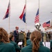 An outdoor memorial or commemorative ceremony is taking place, featuring a speaker at a podium wearing a ceremonial chain of office, suggesting a local official or mayor. Three national flags are displayed on flagpoles — the United Kingdom Union Jack, the Netherlands flag, and the United States flag — along with a military color guard bearing a U.S. Army standard. A band in green uniforms is seated in the foreground with sheet music, suggesting a formal public event likely related to WWII liberation commemoration.