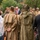 Deputy Mayor of Grave Delivers Remarks at the LT John S Thompson Bridge Ceremony, Operation Market Garden Commemorations 2026 A commemorative or memorial gathering featuring individuals dressed in WWII-era military uniforms and civilian attire. Two figures in the foreground wear period-accurate olive drab military coats and headgear, standing with their backs to the camera. A larger group including people in military reenactment uniforms and formal civilian dress is visible in the background.