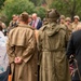 A commemorative or memorial gathering featuring individuals dressed in WWII-era military uniforms and civilian attire. Two figures in the foreground wear period-accurate olive drab military coats and headgear, standing with their backs to the camera. A larger group including people in military reenactment uniforms and formal civilian dress is visible in the background.