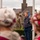 A Scout and WWII Reenactors Participate at the LT John S Thompson Bridge Ceremony, Operation Market Garden Commemorations 2026 A commemorative or memorial ceremony is taking place outdoors, likely in a European town. Scouts wearing distinctive red and white hats are in the foreground, while a young scout with badges on his sleeve stands at attention near a flagpole. In the background, individuals dressed in WWII-era military uniforms (reenactors) are also present, suggesting this may be a liberation or remembrance event.