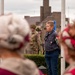A commemorative or memorial ceremony is taking place outdoors, likely in a European town. Scouts wearing distinctive red and white hats are in the foreground, while a young scout with badges on his sleeve stands at attention near a flagpole. In the background, individuals dressed in WWII-era military uniforms (reenactors) are also present, suggesting this may be a liberation or remembrance event.