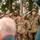 Attendees Render Honors at the LT John S Thompson Bridge Ceremony, Operation Market Garden Commemorations 2026 A group of U.S. Army soldiers in modern MultiCam/OCP uniforms are standing at salute during a formal ceremony, alongside an individual dressed in WWII-era U.S. Army uniform including an M1 helmet and 82nd Airborne Division patch. The setting appears to be an outdoor commemorative event, possibly a liberation or memorial ceremony, suggested by orange decorations visible in the background trees. The mix of modern and historical uniforms indicates this may be a WWII anniversary commemoration.