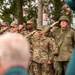 A group of U.S. Army soldiers in modern MultiCam/OCP uniforms are standing at salute during a formal ceremony, alongside an individual dressed in WWII-era U.S. Army uniform including an M1 helmet and 82nd Airborne Division patch. The setting appears to be an outdoor commemorative event, possibly a liberation or memorial ceremony, suggested by orange decorations visible in the background trees. The mix of modern and historical uniforms indicates this may be a WWII anniversary commemoration.