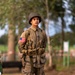 A young man is wearing a detailed WWII-era U.S. Army uniform, including period-accurate equipment and insignia. He is standing on a cobblestone surface outdoors, surrounded by trees, suggesting a commemorative or reenactment event. The uniform includes an American flag patch, M1 helmet with netting, and full combat webbing gear.