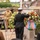 U.S. Navy Attendee Renders Honors at the LT John S Thompson Bridge Ceremony, Operation Market Garden Commemorations 2026 A military memorial ceremony is taking place, featuring a naval officer in dress uniform saluting while placing a floral wreath at a monument. The scene includes re-enactors dressed in WWII-era U.S. Army uniforms alongside vintage military vehicles, suggesting a commemorative event, likely a WWII liberation anniversary. The wreath is decorated with red, white, and blue ribbons consistent with American patriotic colors.