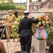 A military memorial ceremony is taking place, featuring a naval officer in dress uniform saluting while placing a floral wreath at a monument. The scene includes re-enactors dressed in WWII-era U.S. Army uniforms alongside vintage military vehicles, suggesting a commemorative event, likely a WWII liberation anniversary. The wreath is decorated with red, white, and blue ribbons consistent with American patriotic colors.