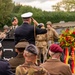 A military memorial ceremony is taking place outdoors, featuring personnel in various uniforms including modern military dress and WWII-era reenactment uniforms. A senior officer in a dark naval uniform with gold braid is saluting at a podium, while a colorful floral wreath with a ribbon bearing German text is prominently displayed. The gathering includes attendees in both contemporary and historical military attire, suggesting a commemorative event likely related to WWII.