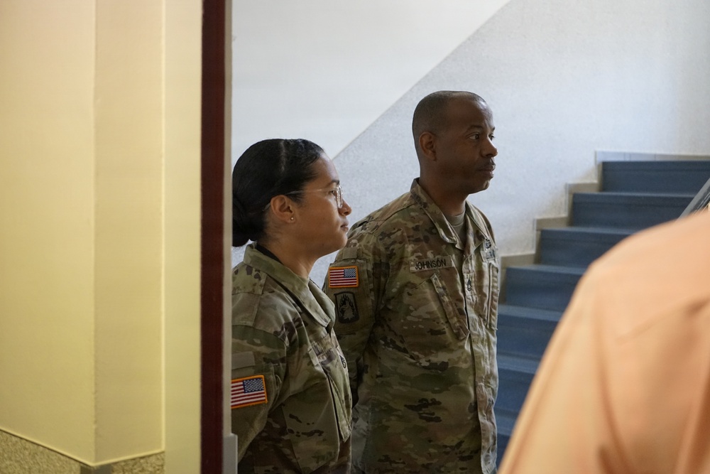 Two U.S. Army soldiers in OCP (Operational Camouflage Pattern) uniforms are standing near a stairwell inside a building, both looking in the same direction. The male soldier's name tape reads 'JOHNSON' and both wear U.S. flag patches on their right shoulders. A third partially visible person in an orange shirt is in the foreground.