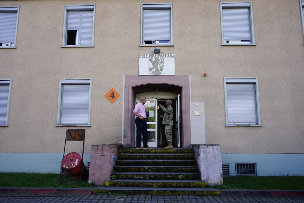 The image shows the entrance of a building labeled 'Frankenberg' with the number 4 marked on an orange diamond sign. Two individuals are standing on the steps at the entrance — one in civilian clothing (pink shirt, dark pants) and one in military camouflage uniform. The building appears to be a European-style institutional or military barracks structure with roller shutters on the windows.
