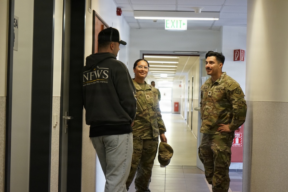 Three individuals are conversing in an indoor corridor of what appears to be a U.S. Army military facility. Two individuals are wearing U.S. Army OCP (Operational Camouflage Pattern) uniforms, while a third person in civilian clothing faces them. The setting is a well-lit institutional hallway with standard safety signage visible.