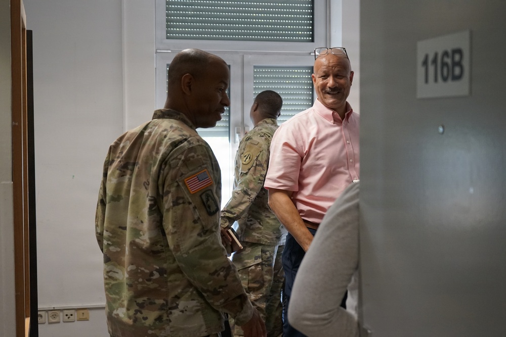 The image shows a group of people in a corridor or hallway near room 116B. Two individuals are wearing U.S. Army camouflage (OCP/MultiCam) uniforms with American flag patches visible, while a third man in a pink polo shirt with glasses on his head is smiling broadly. The setting appears to be an administrative or office building.