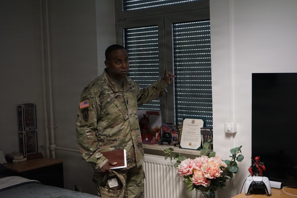 A U.S. Army soldier in MultiCam (OCP) camouflage uniform is standing in what appears to be a barracks or personal quarters room, gesturing and speaking. The soldier wears a U.S. flag patch and a unit patch on the left sleeve, and holds a small notebook or booklet. The room contains personal items including flowers, a PS5 controller, a Deadpool figurine, a framed certificate, and a television.