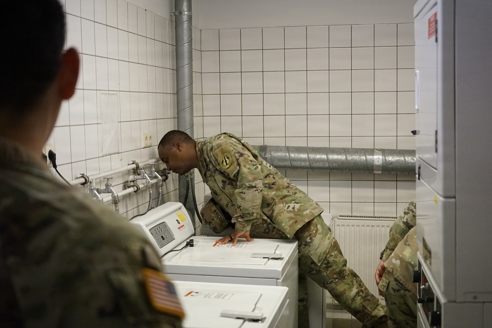 Two or more U.S. Army soldiers in OCP (Operational Camouflage Pattern) uniforms are examining or operating a top-loading washing machine in a tiled utility/laundry room. One soldier is leaning over the machine, appearing to read instructions or inspect the controls. The room features white ceramic tile walls, exposed plumbing, and a ventilation duct.