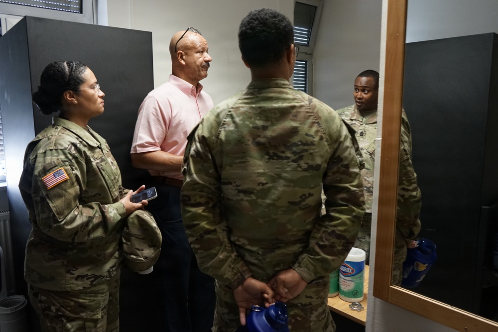Three individuals in U.S. Army OCP (Operational Camouflage Pattern) uniforms are engaged in conversation with a civilian man wearing a pink polo shirt in what appears to be an indoor facility room. A large mirror on the right side of the frame reflects one of the soldiers, providing an additional angle of the scene. Cleaning supplies, including a Clorox wipes container, are visible on a shelf in the background.