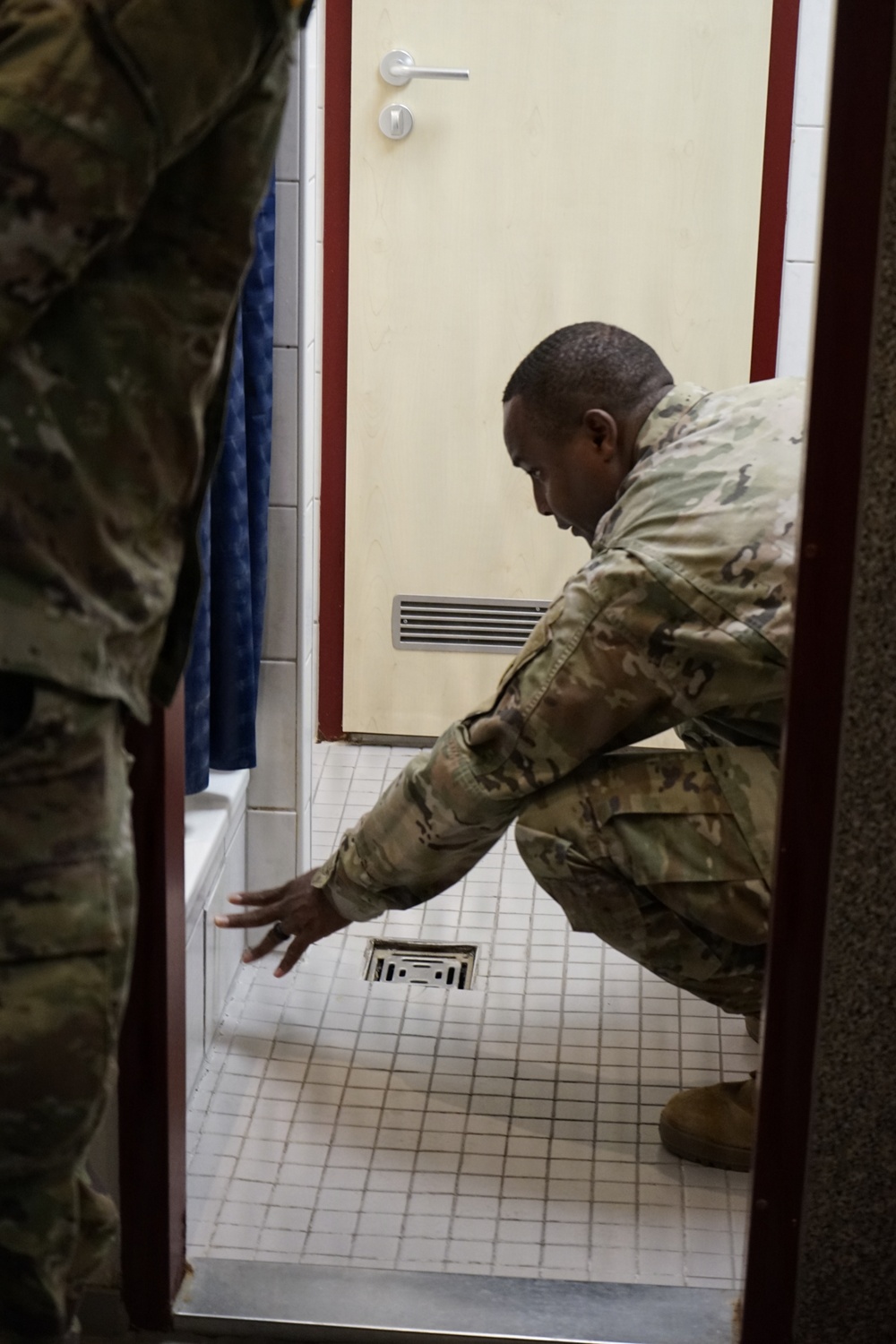 A soldier in MultiCam/OCP camouflage uniform is crouching down inside what appears to be a shower or bathroom facility, pointing toward a floor drain. A second soldier in similar uniform is partially visible on the left side of the frame. The scene suggests an inspection or assessment of the facility's drainage or flooring.