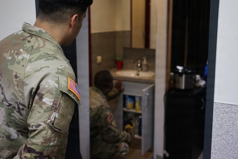 Two U.S. Army soldiers in OCP (Operational Camouflage Pattern) uniforms are visible, with one standing in the foreground and another crouching near a small refrigerator in what appears to be a bathroom or small utility room. The soldier in the foreground has a U.S. flag patch visible on the left sleeve. The setting appears to be a military barracks or quarters.