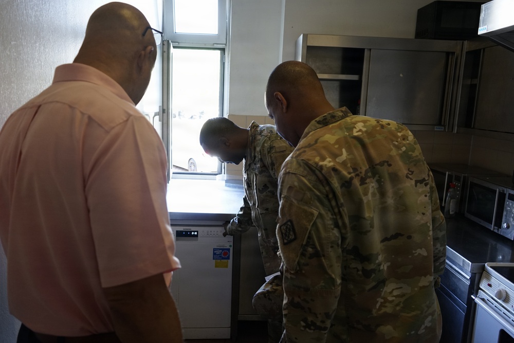 Three individuals are gathered in what appears to be a kitchen or food service area. Two of the individuals are wearing military camouflage uniforms (OCP/MultiCam pattern), while a third person in a pink civilian shirt observes. They appear to be inspecting or examining a dishwasher or similar appliance near a window.