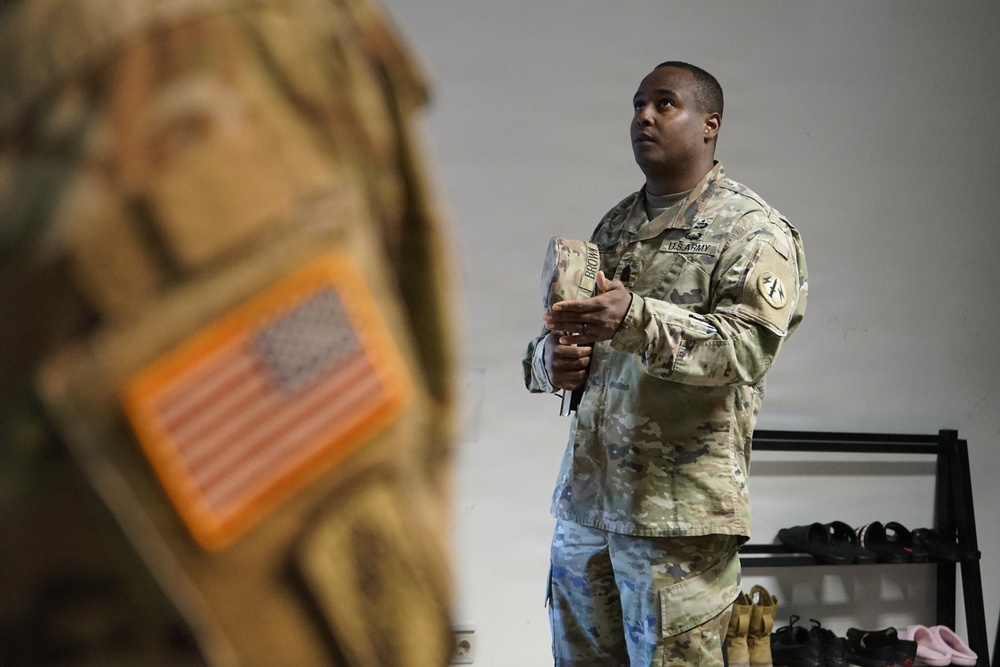 A U.S. Army soldier in MultiCam (OCP) camouflage uniform is standing in what appears to be an equipment room or briefing area, holding a military cap and looking upward or toward something off-frame. A blurred foreground shows another uniform with an orange-bordered U.S. flag patch visible. The setting suggests a pre-deployment preparation, equipment issue, or training environment.