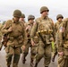 A group of individuals dressed in WWII-era U.S. Army uniforms are marching along a rural road, likely participating in a historical reenactment or commemorative event. They are wearing M1 helmets, olive drab uniforms, and carrying period-accurate equipment including weapons, ammunition pouches, and field gear. American flag patches and unit insignia are visible on their uniforms.