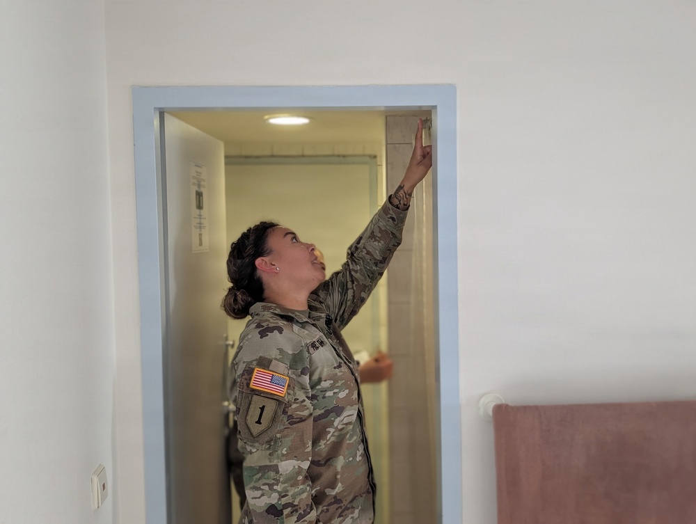 A female U.S. Army soldier in MultiCam/OCP camouflage uniform is inspecting a doorframe or upper door area in what appears to be a building interior. She is pointing upward with one finger toward the top of the door frame while looking up. The setting appears to be a residential or administrative building, possibly during a housing or facility inspection.