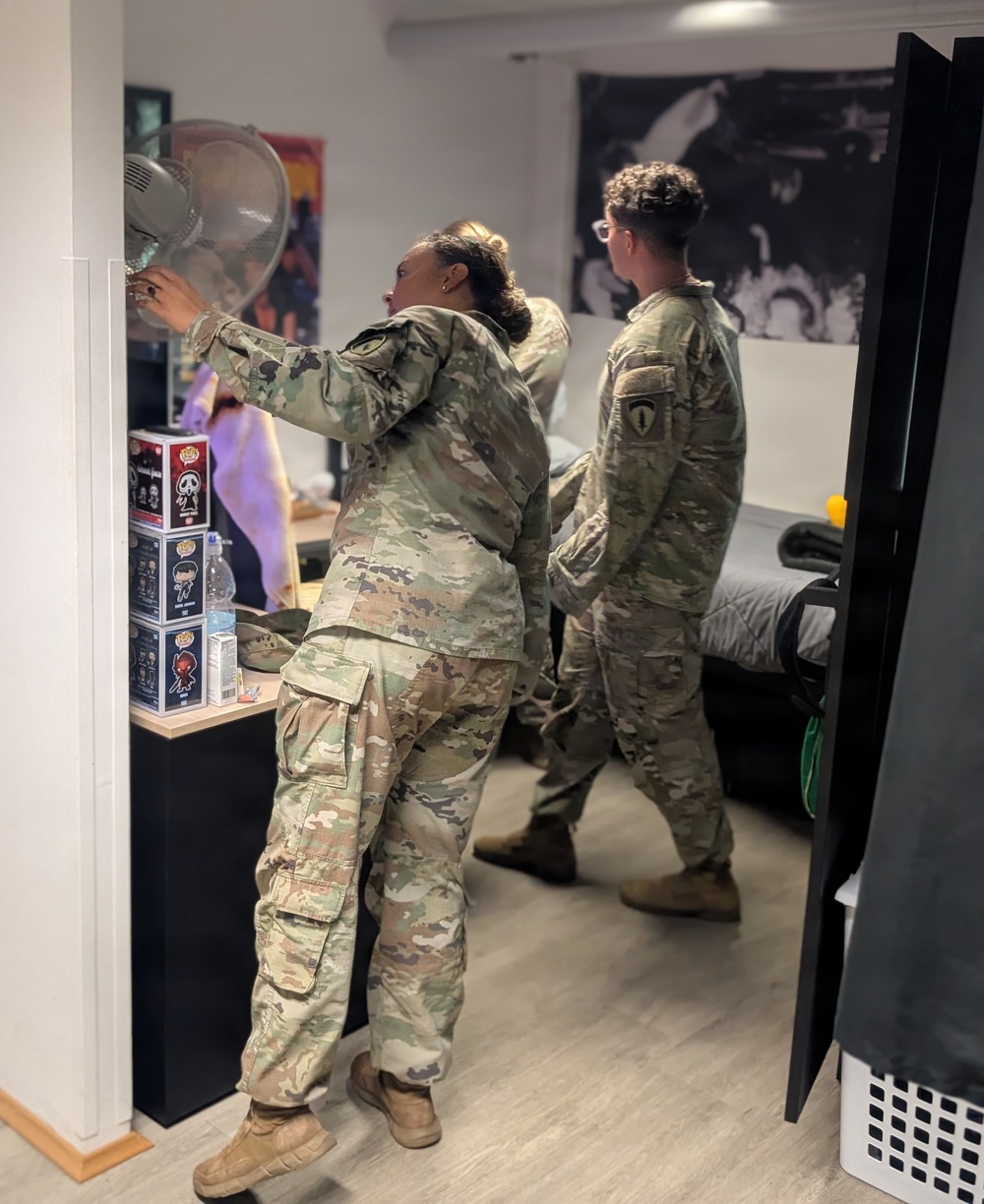 Three individuals wearing military camouflage uniforms (OCP/MultiCam pattern) are inside what appears to be a barracks room or dormitory-style living space. One person in the foreground is reaching toward a standing fan on a desk, while others are moving through the room. The space contains personal items including Funko Pop collectible boxes, a water bottle, and decorative posters on the walls.
