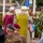 Attendees Render Honors at the LT John S Thompson Bridge Ceremony, Operation Market Garden Commemorations 2026 A memorial or commemoration ceremony is taking place, likely related to World War II, featuring floral wreaths, participants in period military uniforms, and civilian attendees. Two elderly women with white/grey hair stand facing a monument, one wearing a burgundy vest and the other a yellow-green vest. An American flag and a wreath ribbon reading 'Lest We Forget' are visible among the tributes.