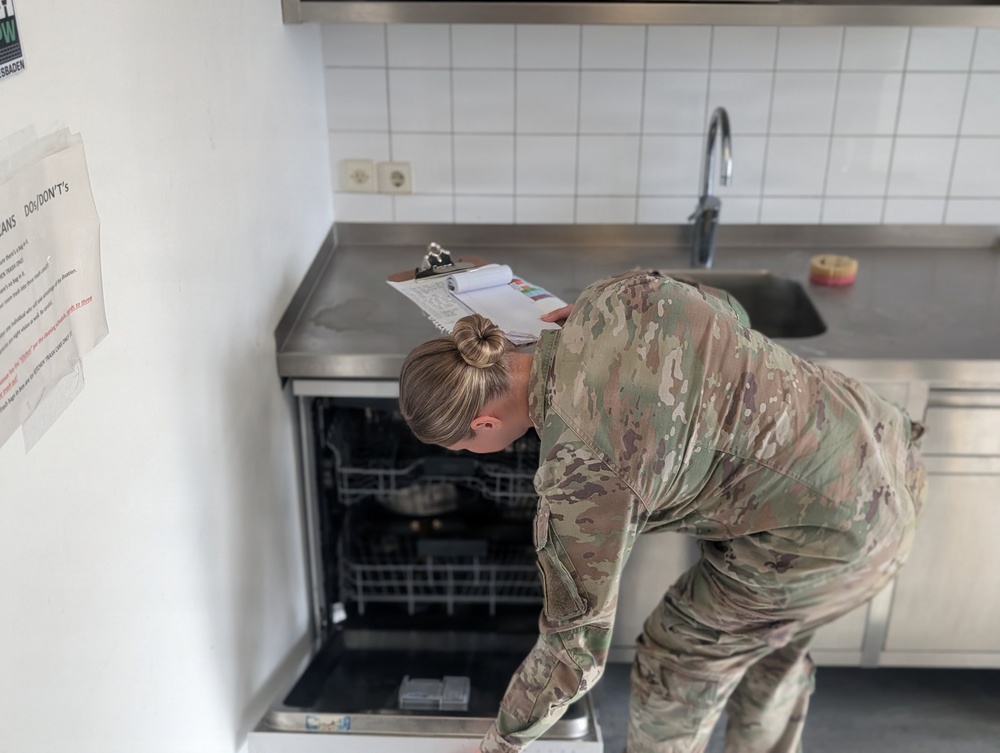A military service member in camouflage uniform is conducting an inspection of a kitchen area, leaning over an open dishwasher while holding a clipboard with documents. The setting appears to be a residential or barracks kitchen, likely on a military installation. A posted notice on the wall labeled 'DO's/DON'Ts' is visible, suggesting this is a formal inspection or compliance check.