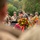 Lt. Col. Travis A. Nedderson Delivers Remarks at the LT John S Thompson Bridge Ceremony, Operation Market Garden Commemorations 2026 A military officer in dress uniform delivers a speech at a podium during what appears to be a memorial or commemoration ceremony. The scene includes personnel wearing WWII-era U.S. Army uniforms and helmets, along with colorful floral wreaths prominently displayed. The image is captured through a shallow depth of field with foreground figures slightly out of focus.
