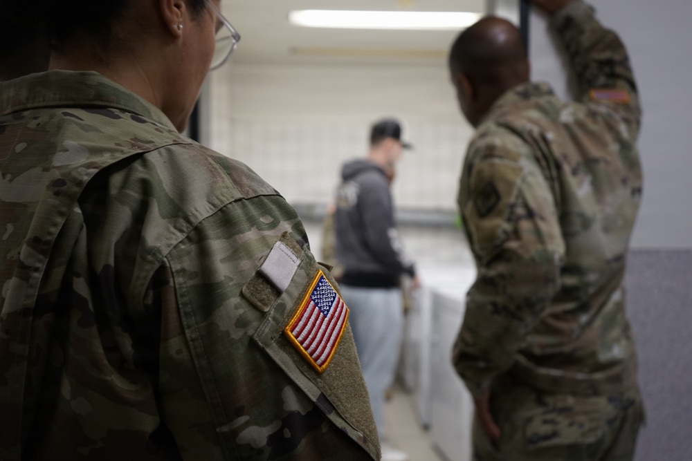 Two U.S. Army soldiers in Operational Camouflage Pattern (OCP) uniforms are observing a civilian individual in an indoor facility. The foreground soldier displays a U.S. flag patch on the right sleeve, consistent with U.S. Army uniform standards. The scene appears to depict a monitoring or oversight activity in what looks like a processing or administrative area.