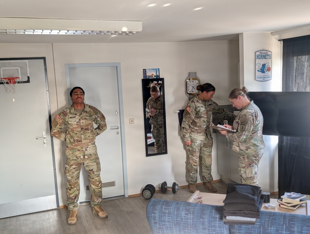 Four U.S. Army soldiers in OCP (Operational Camouflage Pattern) uniforms are inside what appears to be a military barracks room. Two soldiers are writing on clipboards or notepads, suggesting an inspection or inventory is taking place. One soldier stands near the door with hands on hips while another is reflected in a full-length mirror.