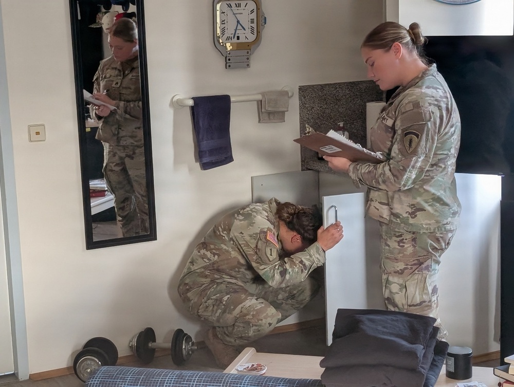 Two female soldiers in US Army OCP (Operational Camouflage Pattern) uniforms are conducting what appears to be a room or barracks inspection. One soldier is crouching down to inspect a low cabinet or furniture piece, while the other stands holding a clipboard and taking notes. A mirror on the left wall reflects one of the soldiers, creating a visible duplicate image.