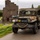 WWII Vehicle Displayed at the LT John S Thompson Bridge Ceremony, Operation Market Garden Commemorations 2026 A restored WWII-era American military truck, identified as a Dodge WC series vehicle, is driving along a narrow road in what appears to be the Netherlands. The vehicle is decorated with period-accurate markings, camouflage netting, and Coca-Cola crates, suggesting participation in a historical military convoy or reenactment event. A brick building bearing the name 'Van Sasse' is visible in the background.