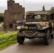 A restored WWII-era American military truck, identified as a Dodge WC series vehicle, is driving along a narrow road in what appears to be the Netherlands. The vehicle is decorated with period-accurate markings, camouflage netting, and Coca-Cola crates, suggesting participation in a historical military convoy or reenactment event. A brick building bearing the name 'Van Sasse' is visible in the background.